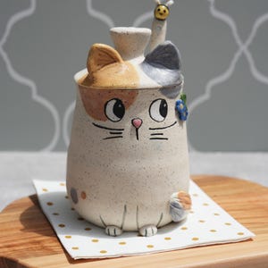 Può includere: Un contenitore in ceramica a forma di gatto con coperchio e manico a forma di ape. Il gatto ha un corpo beige maculato, orecchie arancioni e grigie e un simpatico viso dipinto. È appoggiato su un tovagliolo bianco a pois dorati.