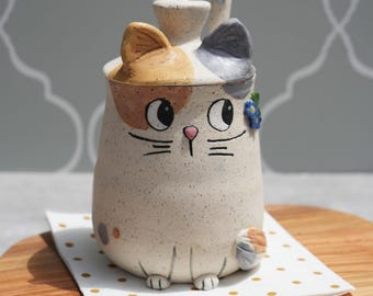 May include: A ceramic cat-shaped container with a lid and a bee-shaped handle. The cat has a speckled beige body, orange and grey ears, and a painted face. It rests on a white napkin with gold dots, on a wooden board.
