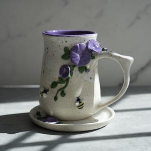 May include: A speckled ceramic mug with a purple rim, adorned with purple flower and green vine details. Two small bees are placed on the mug. The mug sits on a matching saucer.