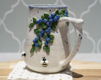 May include: A speckled ceramic mug with a light cream base featuring a raised, three-dimensional design of blue berries and green leaves on the side. The mug also includes small, sculpted bee figures and a sturdy handle with colorful speckles.