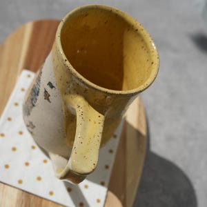 May include: A handmade ceramic mug with a yellow interior and handle, speckled with brown dots. The exterior is off-white with a painted design. The mug sits on a wooden surface with a white and yellow polka dot napkin.