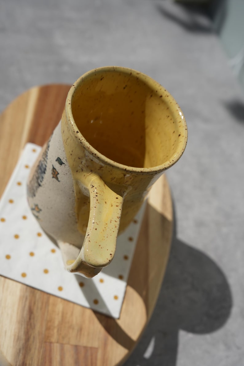May include: A handmade ceramic mug with a yellow interior and handle, speckled with brown dots. The exterior is off-white with a painted design. The mug sits on a wooden surface with a white and yellow polka dot napkin.