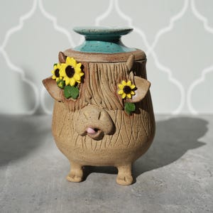 May include: A whimsical ceramic jar with a lid, featuring a Highland cow design. The jar is tan with carved details, a turquoise lid, and adorned with yellow sunflower accents. The jar has a height of approximately 6 inches.
