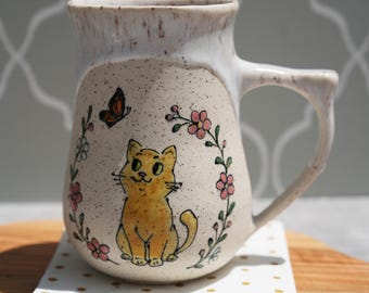 May include: A ceramic mug with a hand-painted design of a yellow cat, a butterfly, and pink flowers. The mug has a cream-coloured base and a light brown rim and handle. It sits on a white napkin with gold dots.