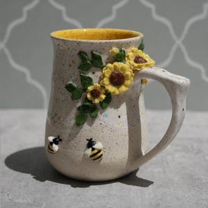 May include: A ceramic mug with a speckled cream glaze and a bright yellow interior. The exterior features three-dimensional sculpted yellow sunflowers with green leaves and two small bumblebees near the base, all attached to the side.