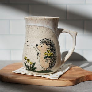 Könnte beinhalten: Beige Keramikbecher mit einer Illustration eines Igels, der eine Pusteblume pustet und gelbe Blumen hält. Der Becher steht auf einem Holzbrett mit einer gepunkteten Serviette. Der Griff hat eine markante, geschwungene Form.