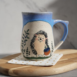 Pode incluir: Caneca de cerâmica com borda e alça azul clara, com ilustração de ouriço-cacheiro pintada à mão. O ouriço-cacheiro segura uma cesta de mirtilos. A caneca está sobre uma pequena toalha de bolinhas em uma tábua de madeira.