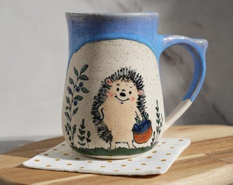 May include: A ceramic mug with a light blue rim and handle, featuring a hand-painted hedgehog illustration. The hedgehog holds a basket of blueberries. The mug is set on a small, dotted napkin on a wooden board.