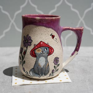Op de afbeelding: Een keramische mok met een grillig ontwerp. De mok toont een grijze kat met een rode paddenstoelhoed, omringd door paarse bloemen en een lieveheersbeestje. De rand en het handvat zijn diep paars. De mok staat op een kleine, goudgestippelde vierkant.