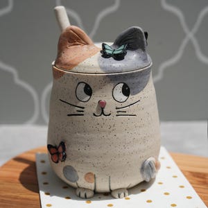 May include: A ceramic cat-shaped container with a lid and spoon. The container is off-white with black speckles, and features a cat face design with a pink nose and whiskers. The lid has a butterfly and the ears are brown and grey. The container sits on a dotted napkin.