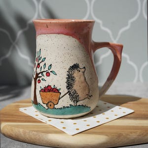 Pode incluir: Caneca de cerâmica pintada à mão com um design caprichoso. A caneca tem um fundo creme salpicado com uma borda e uma alça rosa. Um ouriço de desenho animado puxa uma carroça de maçãs perto de uma árvore. A caneca está em uma tábua de madeira com um guardanapo pontilhado.