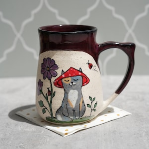 May include: A ceramic mug with a burgundy rim and handle. The mug features a hand-painted design of a cat wearing a red mushroom hat, sitting near purple flowers and a ladybug. The mug is on a small square napkin with gold dots.