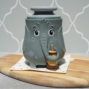 May include: A speckled gray ceramic canister shaped like an elephant with a lid. The elephant has painted eyes, eyelashes, and a trunk holding a small brown and yellow brush. The canister features a smooth finish with subtle speckles throughout.