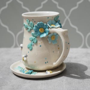 Op de afbeelding: Crèmekleurige keramische mok en schotelset, versierd met turquoise bloemen en bladeren. De mok heeft een handvat en is versierd met kleine bijen. De schotel weerspiegelt het bloemendesign. Een charmant, handgemaakt item.