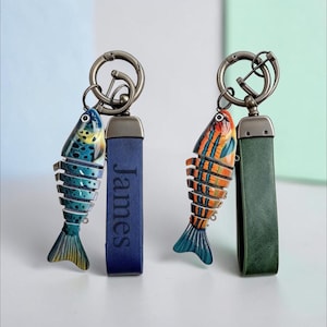 Könnte beinhalten: Zwei Schlüsselanhänger mit beweglichen Fisch-Anhängern an Metallringen. Ein Schlüsselanhänger hat einen blauen Fisch mit schwarzen Punkten und ein blaues Lederband mit dem eingeprägten Namen James. Der andere hat einen orange-blau gestreiften Fisch.