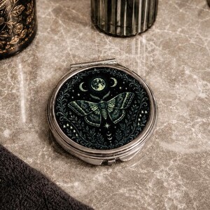 May include: A round silver compact mirror featuring a dark green and black illustration of a moth centered beneath a moon and stars. The design is surrounded by a detailed botanical border of leaves and vines, creating a cohesive circular graphic.