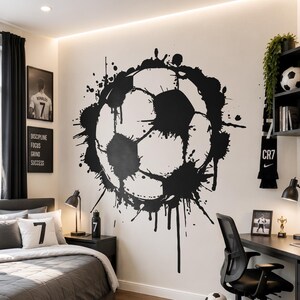 Könnte beinhalten: Ein großer Wandaufkleber in Form eines Fußballes mit schwarzem Tintenspritzer-Effekt, zentriert an einer weißen Wand in einem Schlafzimmer. Der Raum enthält ein graues Bett mit Kissen, die mit der Zahl 7 markiert sind, einen Schreibtisch und Rucksack.