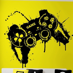 Könnte beinhalten: Schwarzer und weißer Videospiel-Controller-Aufkleber an einer gelben Wand mit einem weißen Sofa und schwarzen, weißen und gelben Kissen. Der Aufkleber trägt das Wort "GAME".
