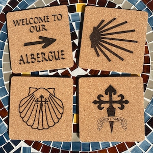 Puede incluir: Cuatro posavasos de corcho con diferentes diseños relacionados con la peregrinación del Camino de Santiago. El primer posavasos dice "Bienvenido a nuestro Albergue" con una flecha apuntando hacia la derecha. El segundo posavasos presenta el símbolo de la concha de vieira. El tercer posavasos tiene una concha de vieira con una cruz dentro. El cuarto posavasos tiene una cruz con las palabras "Buen Camino" debajo.