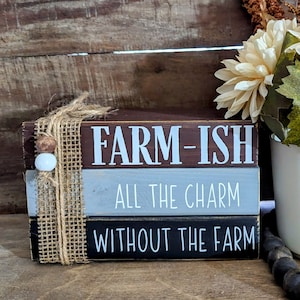 May include: Stacked wooden blocks with the text "FARM-ISH", "ALL THE CHARM", and "WITHOUT THE FARM" in white paint. The blocks are decorated with burlap ribbon and white beads. The blocks are sitting on a wooden surface with a white ceramic pot with a bouquet of cream-colored flowers.
