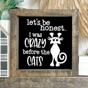 May include: A black wooden sign with white text that reads "let's be honest... I was crazy before the cats". The sign features a white silhouette of a cat with its ears up and a tail curled around its body. The sign is sitting on a shelf with a white heart-shaped object and a white box with the text "Blifftin House" on it.