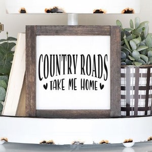 May include: A white tiered tray with a wooden sign that reads "COUNTRY ROADS TAKE ME HOME" with hearts on either side of the text. The tray is decorated with greenery, books, and wooden beads.