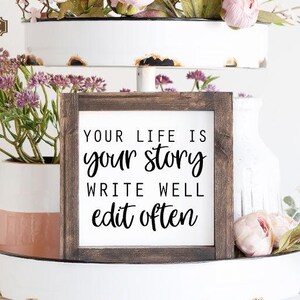 May include: A wooden sign with the text "Your life is your story write well edit often" on a white tiered tray with pink flowers and greenery.