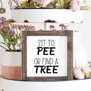 May include: A rustic wooden sign with the text "Sit to Pee or Find a Tree" in black lettering. The sign is displayed on a white tiered tray with pink flowers.