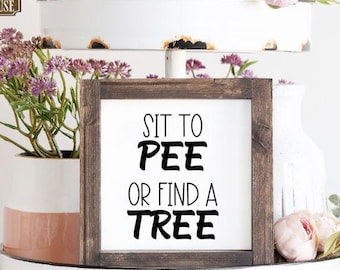 May include: A rustic wooden sign with the text "Sit to Pee or Find a Tree" in black lettering. The sign is displayed on a white tiered tray with pink flowers.