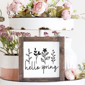 May include: A white tiered tray with a wooden sign that says "hello spring" in black lettering. The sign is decorated with a black silhouette of five stylised plants. The tray is decorated with pink flowers and greenery.