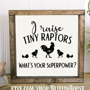 May include: A wooden sign with a white background and black text that reads "I raise tiny raptors. What's your superpower?"  The sign features a black silhouette of a hen and three chicks, with one chick hatching from an egg. The sign is framed in a dark brown wood frame.