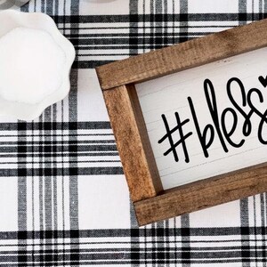 May include: A wooden sign with a white background and black lettering that reads "#blessed" with a small heart symbol. The sign is sitting on a black and white checked surface.