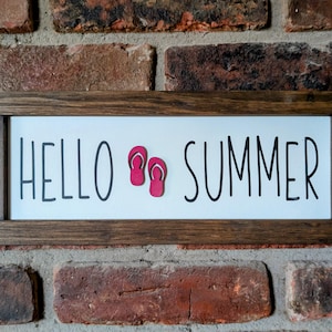May include: A wooden sign with a white background and the text "HELLO SUMMER" in black lettering. Two red flip-flop cutouts are placed between the words.