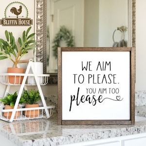 May include: A brown wooden framed sign with white background and black text that reads "We aim to please. You aim too please." with a heart symbol at the end of the text.