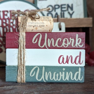May include: Stacked wooden blocks with the words "Uncork and Unwind" painted in white and red lettering on a red, white, and green background. A cork with twine is wrapped around the blocks.
