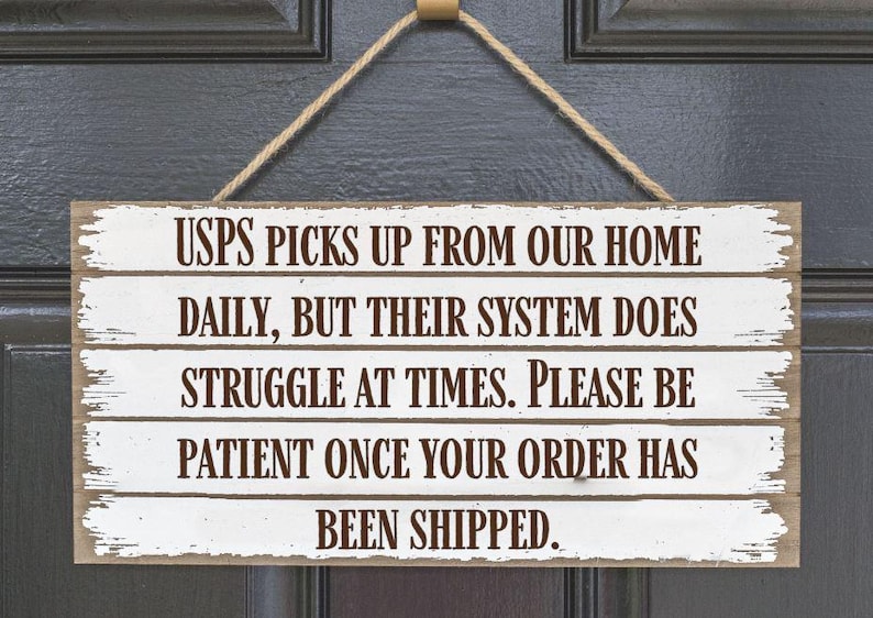 May include: A rectangular wooden sign with white painted planks and brown text. The sign reads: "USPS PICKS UP FROM OUR HOME DAILY, BUT THEIR SYSTEM DOES STRUGGLE AT TIMES. PLEASE BE PATIENT ONCE YOUR ORDER HAS BEEN SHIPPED."