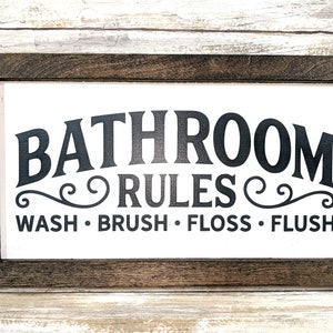 K&ouml;nnte beinhalten: Ein wei&szlig;es Holzschild mit schwarzem Rahmen und dem Text "Bathroom Rules" in einer dekorativen Schriftart. Unter dem Text steht "Wash. Brush. Floss. Flush."