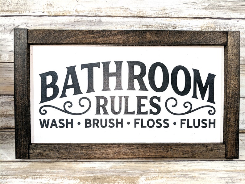 K&ouml;nnte beinhalten: Ein wei&szlig;es Holzschild mit schwarzem Rahmen und dem Text "Bathroom Rules" in einer dekorativen Schriftart. Unter dem Text steht "Wash. Brush. Floss. Flush."
