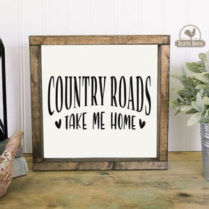 May include: A rustic wooden sign with the text "COUNTRY ROADS TAKE ME HOME" in black lettering with two small hearts on either side of the text. The sign is on a white wood background.