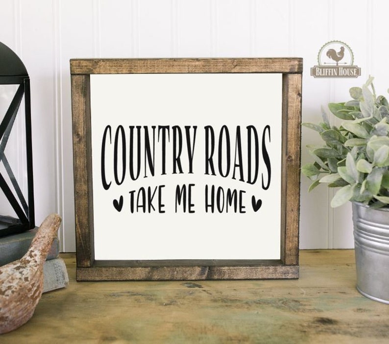 May include: A rustic wooden sign with the text "COUNTRY ROADS TAKE ME HOME" in black lettering with two small hearts on either side of the text. The sign is on a white wood background.