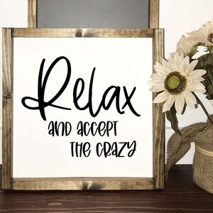 May include: A white wooden sign with black lettering that reads "Relax and accept the crazy". The sign is framed in a rustic brown wood frame.