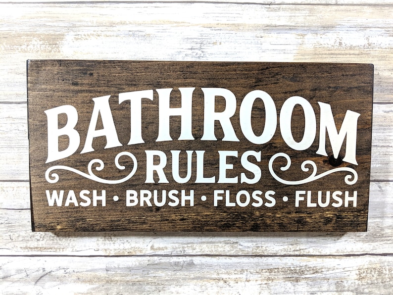 K&ouml;nnte beinhalten: Ein braunes Holzschild mit wei&szlig;em Text, der "Bathroom Rules" lautet, mit dekorativen Schn&ouml;rkeln auf beiden Seiten des Wortes "Rules". Unter dem Text steht "Wash. Brush. Floss. Flush."