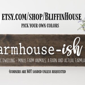 以下が含まれることがあります： 「farmhouse-ish」という文字が白く塗られた素朴な木製の看板。看板には「(N). A rustic dwelling - minus farm animals, a barn and actual farmland」と「(corners are not sanded unless requested」という文字も書かれている。