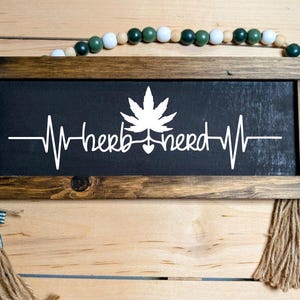May include: A wooden sign with a black background and white text that reads "herb nerd" with a stylized heart and a marijuana leaf. The sign has a brown frame and is hanging from a string of green and white beads.