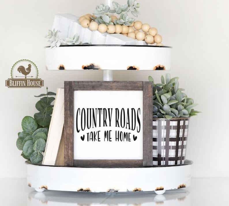 May include: A white tiered tray with a wooden sign that reads "COUNTRY ROADS TAKE ME HOME" with hearts on either side of the text. The tray is decorated with greenery, books, and wooden beads.
