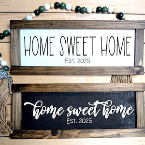 May include: Two framed wooden signs with the text "HOME SWEET HOME EST. 2025" in white on a white background and in white script on a black background. A beaded garland with green, white, and brown beads is draped across the top.