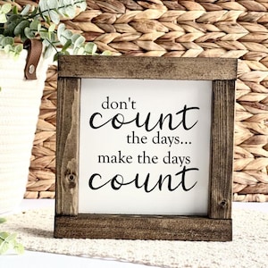 May include: A wooden framed sign with the text "don't count the days... make the days count" in black lettering on a white background.