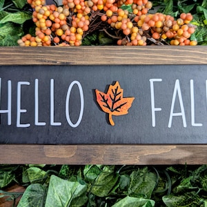 May include: A wooden sign with a black chalkboard insert that reads "HELLO FALL" in white lettering. A small orange and red maple leaf is centered between the words.