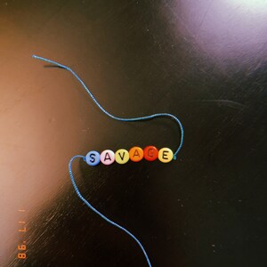 May include: A beaded bracelet spelling out the word "SAVAGE" in colorful letter beads. The beads are strung on a blue cord, with the ends of the cord visible. The bracelet is set against a dark background.