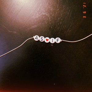 May include: A handmade bracelet with white letter beads spelling "ASIF" and a red heart bead. The bracelet is strung on a thin white cord, set against a dark, textured surface. The date "12. 6 '21" is in the upper right corner.